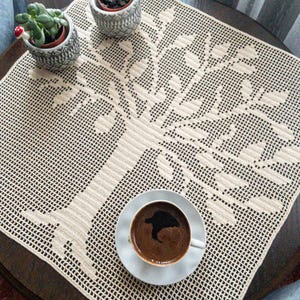 Puede incluir: Un mantel de crochet blanco con un diseño de árbol. El mantel está sobre una mesa de madera con una taza de café y un platillo.