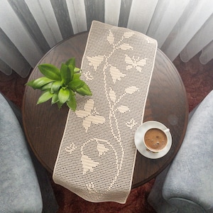 Puede incluir: Un camino de mesa de ganchillo con un diseño floral se encuentra sobre una mesa de madera con una taza de café en un platillo. Una planta verde está en el fondo.