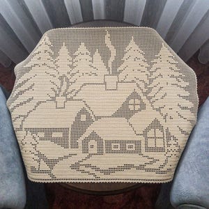 Peut inclure: Une nappe en crochet beige avec un motif de deux maisons et d'arbres. La nappe est sur une table brune.