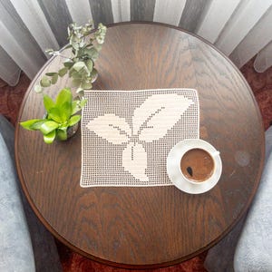 May include: A white crocheted doily with a leaf design sits on a dark wood table with a cup of coffee and a saucer.