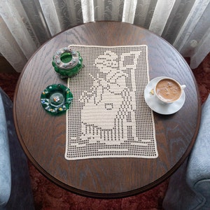 以下が含まれることがあります： 暗い木製のテーブルの上に、花柄のクロシェレースのテーブルクロスが置かれています。テーブルクロスの上には、緑色のガラス製の小箱が2つと、白いソーサーの上に置かれたコーヒーカップがあります。