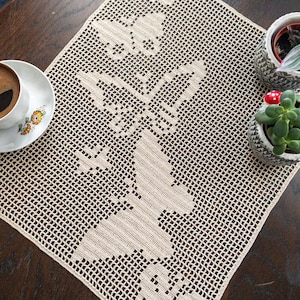 May include: A beige crocheted table runner with a delicate floral design. The runner is laid on a wooden table with two small potted plants.