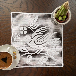 Puede incluir: Doily de crochet blanco con un diseño floral sobre una mesa de madera con una taza de café y un cactus en maceta.