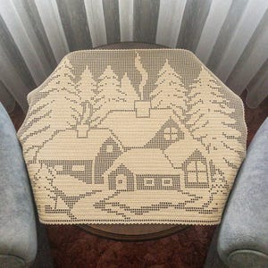 Puede incluir: Cubremesa de ganchillo color crema con una escena invernal. El diseño incluye una casa, árboles y un borde decorativo. El cubremesa está colocado sobre una mesa redonda marrón, con una silla gris a cada lado.
