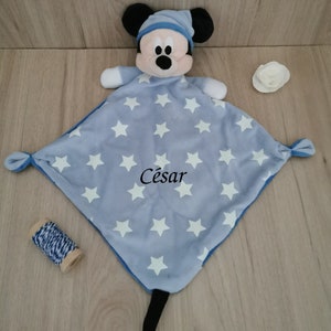 Peut inclure: Une couverture pour bébé bleu et blanche avec une tête de Mickey Mouse et le nom "César" brodé dessus.