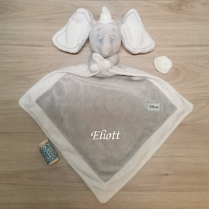 Peut inclure: Une couverture de sécurité en peluche éléphant gris et blanc avec le nom "Eliott" brodé dessus. La couverture a une bordure blanche et un logo Disney.