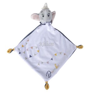 Peut inclure: Une couverture de sécurité pour bébé blanche avec une tête de peluche Dumbo. La couverture a une bordure bleu foncé et un motif de guirlande de triangles colorés. Le nom "Kilian" est brodé sur la couverture, avec le mot "Dumbo" en dessous.