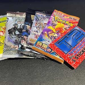 Könnte beinhalten: Eine Sammlung von Sammelkarten- und Aufkleberpackungen. Die Packungen zeigen verschiedene Designs, darunter eine grüne Packung mit einem Hirsch, eine Packung mit Robotern, eine Packung mit Anime-Charakteren und eine rote Aufkleberpackung mit dem Text "TERMINATOR 2".