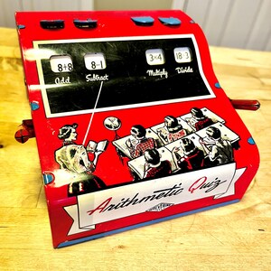 May include: A vintage red metal Arithmetic Quiz toy with blue accents. The toy features math problems for addition, subtraction, multiplication, and division. A graphic shows students in a classroom. The toy is labeled "Wolverine".