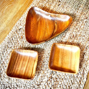 May include: Three wooden serving dishes. One is a triangular shape, and two are square. The dishes are made of a light brown wood with visible grain. They are arranged on a woven, beige placemat.