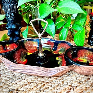 May include: A vintage serving tray with two bowls and a center handle. The tray has a brown, wood-like texture with black, red, and yellow accents. Two tiki statues and a plant are in the background.