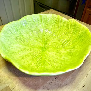 May include: A bright green ceramic serving bowl with a leaf design. The bowl has a scalloped edge and detailed veining patterns, creating a textured appearance. The bowl is sitting on a wooden surface.