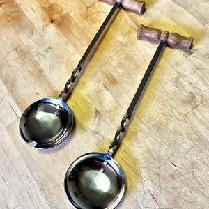 May include: Two silver serving spoons with twisted handles and wooden T-shaped handles. The spoons have a round bowl shape with a small hole in the center.