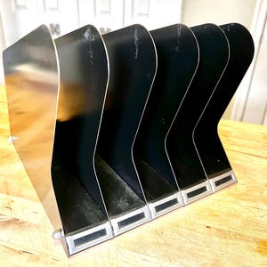 May include: A metal desk organizer with six black compartments. The organizer has a silver-colored side and a rectangular base with a silver trim. The compartments are angled and designed to hold documents or files.