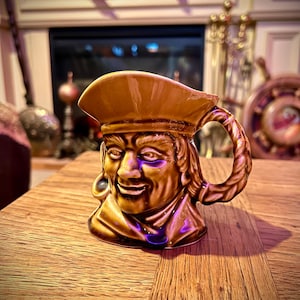 May include: A brown ceramic mug shaped like a pirate's head with a wide brimmed hat. The mug has a handle shaped like a rope. The mug is sitting on a wooden table.