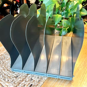 May include: A gray metal desk organizer with five curved dividers. The organizer is made of metal and has a light blue base. The dividers are shaped like stylized leaves. The organizer sits on a wooden table.