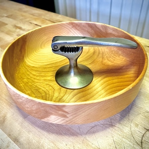 May include: A wooden bowl with a natural finish, showcasing a warm, light brown color. A metal nutcracker with a silver handle and a brass base rests inside the bowl. The bowl's interior displays a beautiful wood grain pattern.
