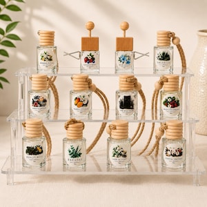Op de afbeelding: Een collectie van twaalf kleine glazen autodiffuserflesjes met houten doppen en ophangkoorden, gerangschikt op een transparante acryl displaystandaard met drie niveaus. Elk flesje heeft een uniek rond etiket met botanische illustraties.