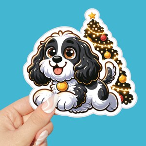 May include: A sticker featuring a cartoon dog with black and white fur, a gold collar, and a happy expression. Behind the dog is a Christmas tree decorated with lights and ornaments. The sticker is held against a blue background.