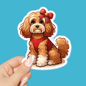 May include: A sticker featuring a cartoon dog with brown fur, white paws, and a red bow. The dog wears a red harness and sits on a sandy surface against a light blue background. The sticker is held by a hand with manicured nails.
