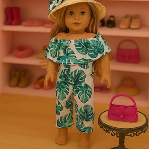 Puede incluir: Muñeca con un mono blanco con estampado de hojas tropicales verdes y un sombrero de paja con una cinta azul. Un pequeño bolso rosa está sobre una mesa decorativa. La muñeca está en una habitación rosa.