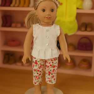 Puede incluir: Una muñeca vestida con una blusa blanca con mangas con volantes y tres botones, combinada con pantalones capri con estampado floral. La muñeca tiene el pelo rubio en una cola de caballo y está de pie sobre una base circular blanca. El fondo incluye una estantería con accesorios para muñecas.