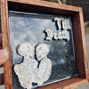 Op de afbeelding: Een houten frame met een glazen voorzijde, met twee skeletten die elkaar omhelzen. De skeletten zijn met een laser uit hout gesneden en wit geverfd. De achtergrond is donkergrijs met een gespikkelde textuur. De tekst "Till Death" is in witte cursieve letters bovenaan het frame geschreven.