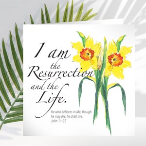May include: A white greeting card with the text "I am the Resurrection and the Life." features a watercolour painting of two yellow daffodils with orange centres. The card also includes the Bible verse John 11:25.