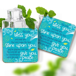 May include: Two square coasters with a turquoise background and white and yellow accents. The coasters feature the text "The Lord bless you and keep you, the Lord make His face shine upon you and give you peace." Mint sprigs and ice cubes are in the background.