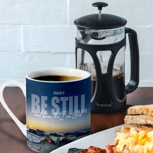May include: A white ceramic mug with a blue background and a sunrise scene. The mug has the text "Be Still and know that I am God." and "Psalm 46:10" printed on it.