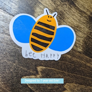 Puede incluir: Una pegatina blanca con una abeja de dibujos animados con rayas negras y alas azules. El texto "bee happy" está escrito debajo de la abeja.