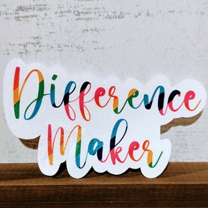 May include: White sign with the words "Difference Maker" in colorful, rainbow-striped letters.