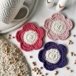 Könnte beinhalten: Drei gehäkelte Untersetzer in Form von Blumen in Pink, Lila und Rot mit einem weißen Zentrum. Die Untersetzer liegen auf einer weißen Oberfläche mit kleinen braunen Bohnen, die um sie herum verstreut sind.