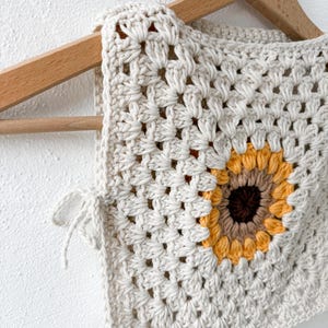 Könnte beinhalten: Cremefarbene Häkelweste mit Sonnenblumenmuster. Die Weste ist aus einem quadratischen Granny-Stichmuster gefertigt. Die Sonnenblume mit gelben, orangefarbenen und braunen Blütenblättern ist mittig platziert. Die Weste hängt an einem Holzkleiderbügel.