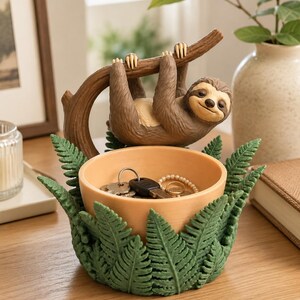 Peut inclure: Un bol de rangement décoratif représentant un paresseux brun suspendu à une branche courbée au-dessus d'un bassin couleur pêche. L'extérieur du bol est orné de feuilles de fougère vertes sculptées. Le bassin contient des clés et une bague.