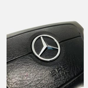 May include: Close-up of a black car steering wheel featuring the Mercedes-Benz logo. The logo is a silver emblem with a three-pointed star within a circle. The letters "SRS AIRBAG" are visible.