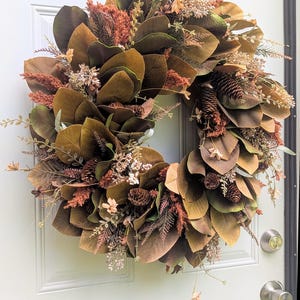 May include: A circular wreath featuring large, dark brown magnolia leaves interspersed with rust-colored plumes, small pinecones, and delicate sprigs of dried foliage. The arrangement creates a dense, layered texture in earthy tones of brown, bronze, and muted orange.