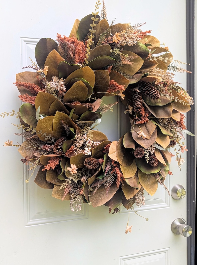 May include: A circular wreath featuring large, dark brown magnolia leaves interspersed with rust-colored plumes, small pinecones, and delicate sprigs of dried foliage. The arrangement creates a dense, layered texture in earthy tones of brown, bronze, and muted orange.