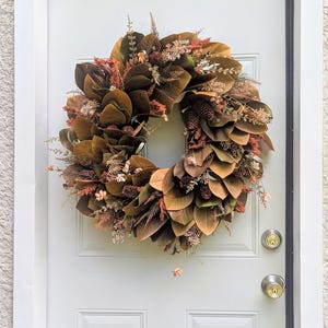 May include: A circular wreath featuring dark brown magnolia leaves, interspersed with small pinecones, dried rust-colored foliage, and delicate beige sprigs. The arrangement creates a dense, layered texture with earthy autumn tones.