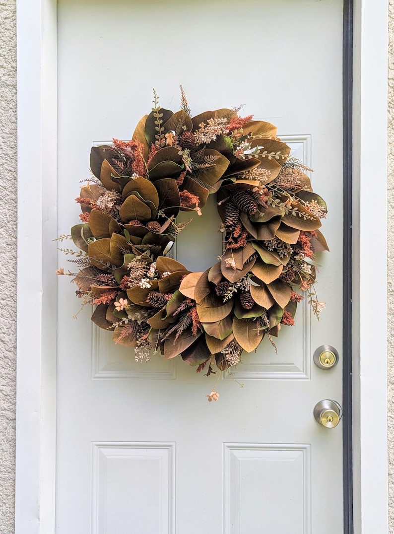 May include: A circular wreath featuring dark brown magnolia leaves, interspersed with small pinecones, dried rust-colored foliage, and delicate beige sprigs. The arrangement creates a dense, layered texture with earthy autumn tones.