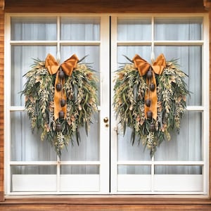 May include: Two teardrop-shaped door wreaths featuring a mix of green foliage, dried grasses, and small white flowers. Each wreath is accented with a large, burnt orange satin bow and several dark brown acorn-shaped ornaments hanging vertically.
