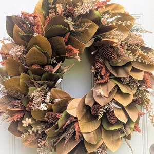 May include: A circular wreath featuring large, broad brown and green magnolia leaves interspersed with textured rust-colored foliage, small white berry clusters, and several brown pinecones arranged in a dense, layered botanical display.