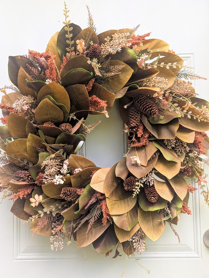 May include: A circular wreath featuring large, broad brown and green magnolia leaves interspersed with textured rust-colored foliage, small white berry clusters, and several brown pinecones arranged in a dense, layered botanical display.