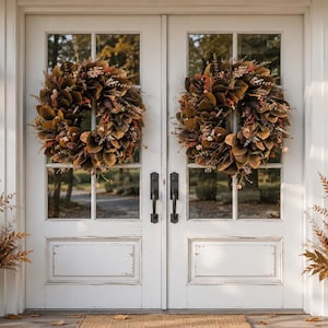 May include: Two identical autumn wreaths featuring brown magnolia leaves, pinecones, and dried foliage are mounted on white double glass doors. Two black metal lanterns flank the doors, and two galvanized metal buckets hold dried plants.