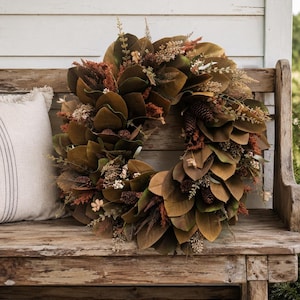 May include: A circular wreath composed of dark brown magnolia leaves, interspersed with small pinecones, dried rust-colored foliage, and sprigs of cream-colored berries. The arrangement features a dense, layered texture with earthy tones.