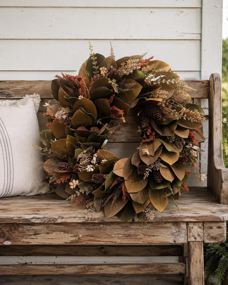 May include: A circular wreath composed of dark brown magnolia leaves, interspersed with small pinecones, dried rust-colored foliage, and sprigs of cream-colored berries. The arrangement features a dense, layered texture with earthy tones.