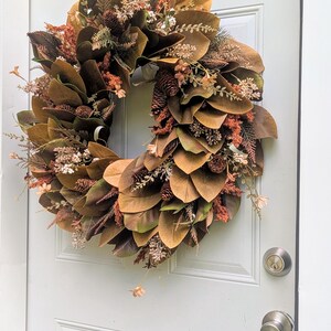 May include: A circular wreath featuring brown magnolia leaves, small pinecones, and dried floral accents in shades of rust, cream, and muted green. The arrangement is densely layered with foliage and botanical elements, creating a textured appearance.