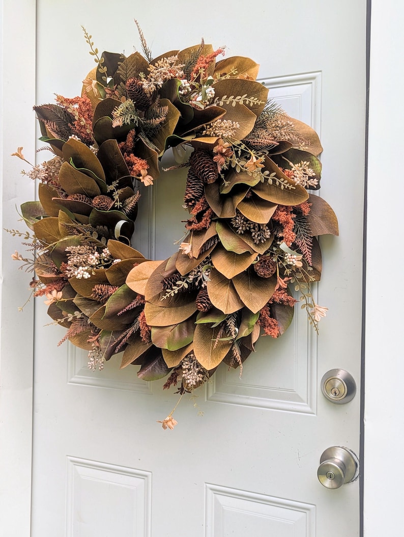 May include: A circular wreath featuring brown magnolia leaves, small pinecones, and dried floral accents in shades of rust, cream, and muted green. The arrangement is densely layered with foliage and botanical elements, creating a textured appearance.