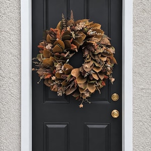 May include: A circular wreath composed of brown magnolia leaves, dried orange and tan foliage, and small pinecones. The wreath is mounted on a solid black door featuring two brass-colored round doorknobs and a white door frame trim.