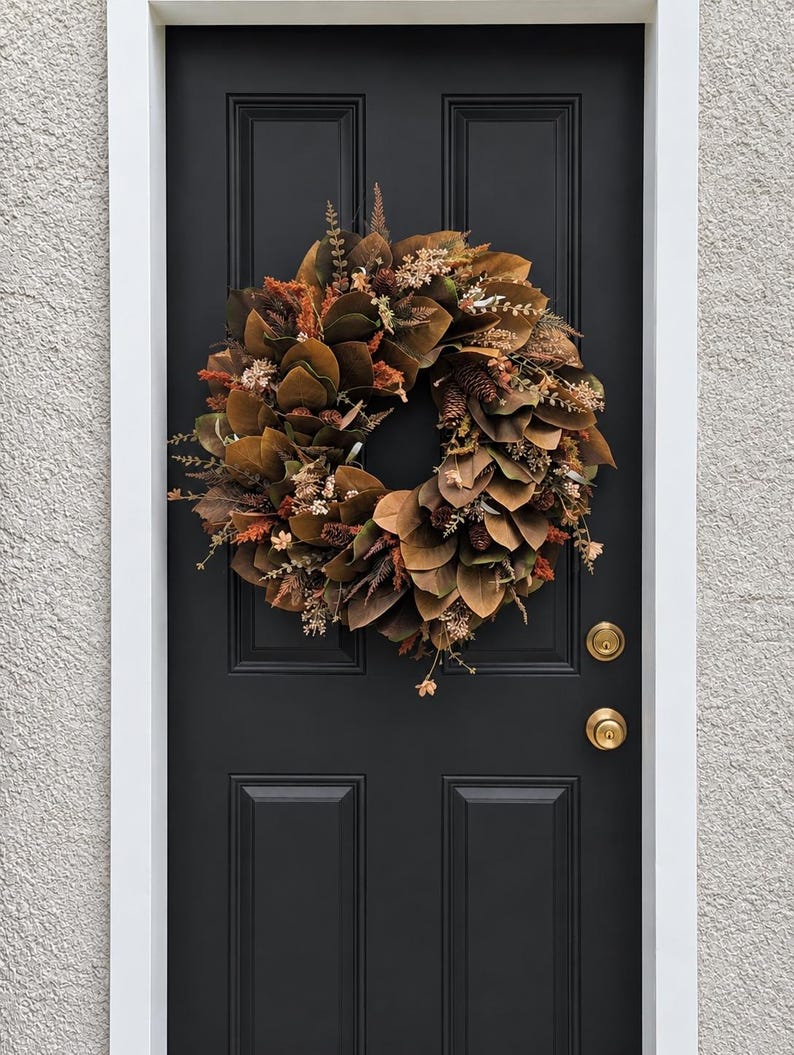 May include: A circular wreath composed of brown magnolia leaves, dried orange and tan foliage, and small pinecones. The wreath is mounted on a solid black door featuring two brass-colored round doorknobs and a white door frame trim.